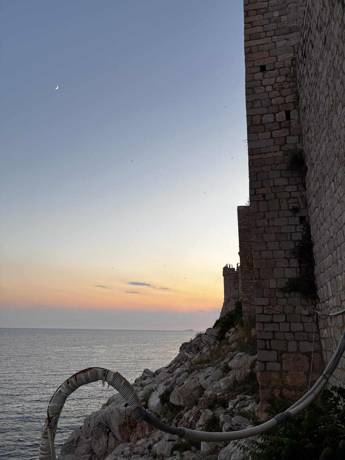 Dubrovniks mächtige Stadtmauer über der Adria im Sonnenuntergang, mit schmaler Mondsichel am Himmel